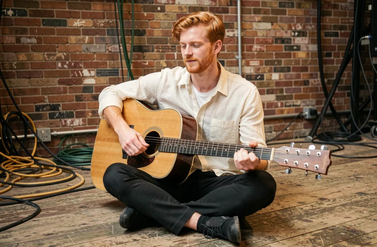 Chanteur roux britannique assis avec une guitare acoustique en coulisses d'une salle de concert, ambiance documentaire authentique